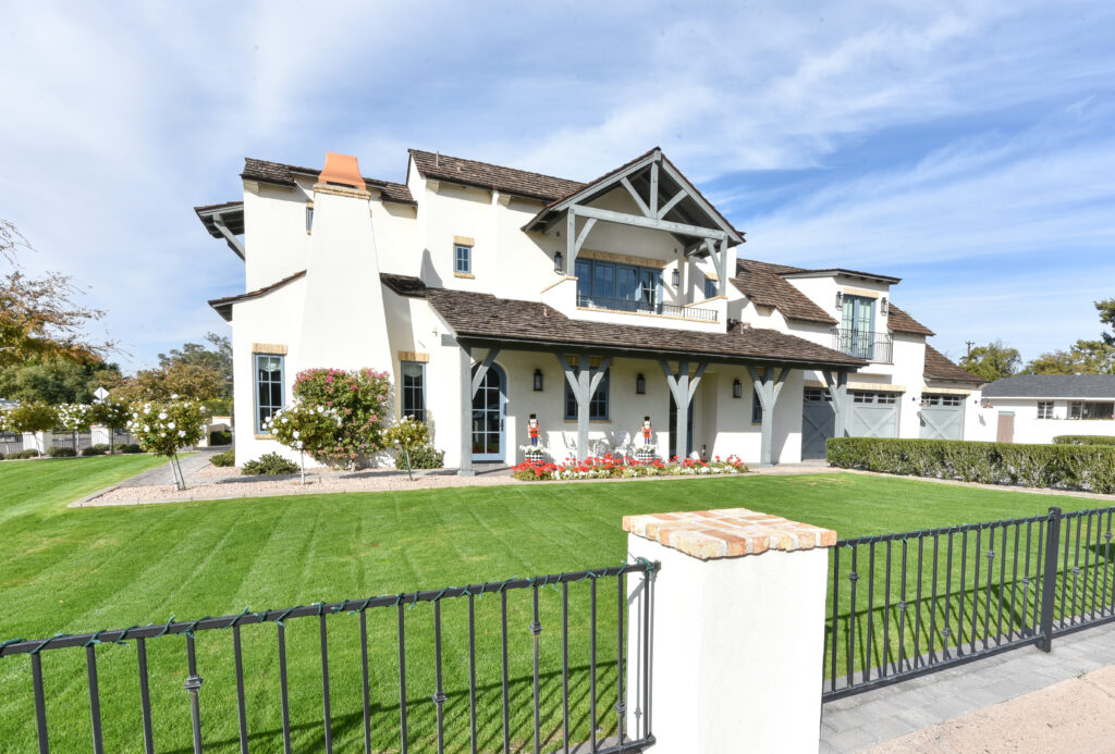 Side profile of the home with a ground-floor porch on grey timber posts, an upper-level gabled balcony, a tapered white stucco chimney, red and white flowerbeds and a green lawn behind the black iron fence