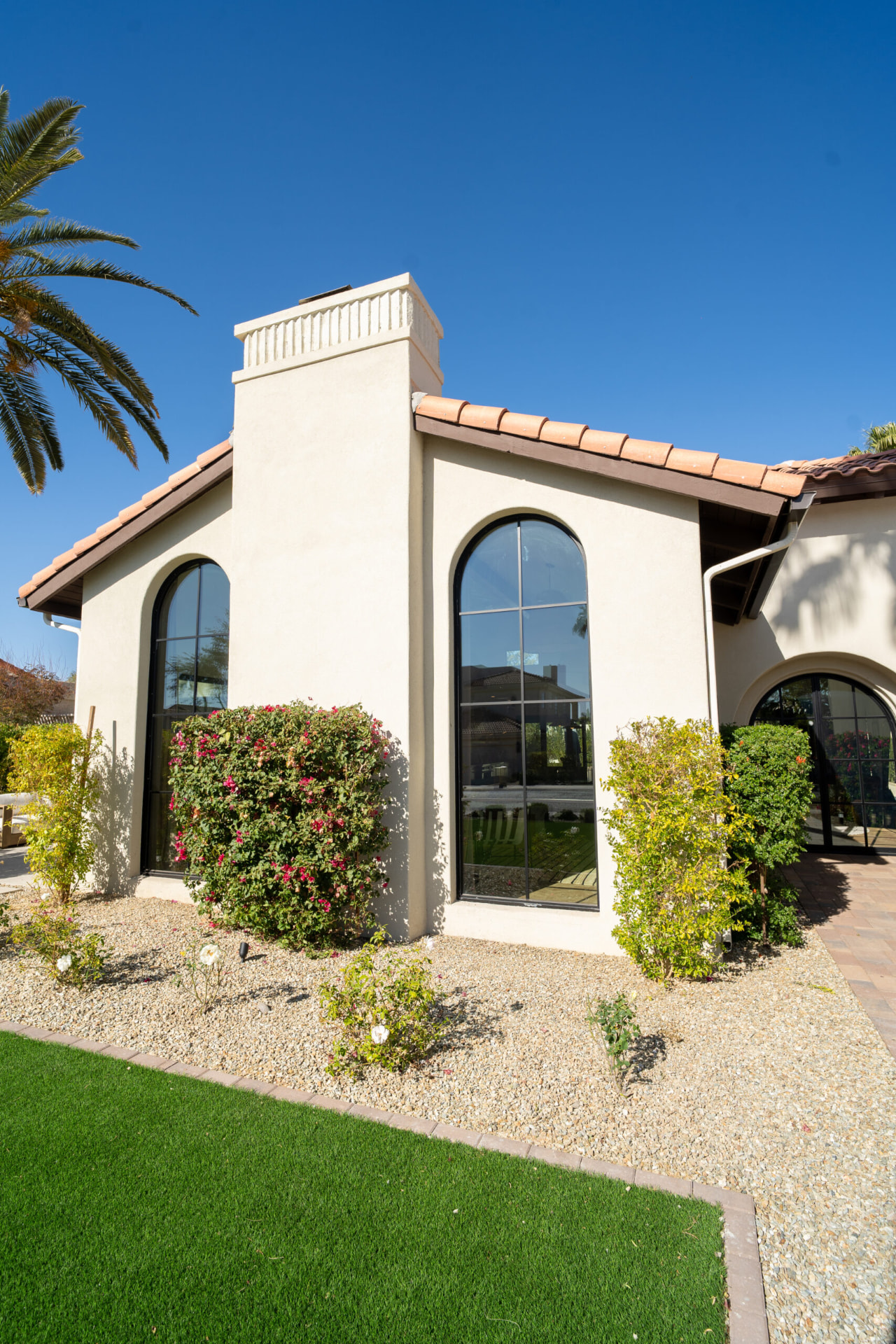 Exterior of the cream stucco home with a prominent chimney, terracotta roof tiles and two tall black metal-framed arched windows above a gravel yard with flowering shrubs
