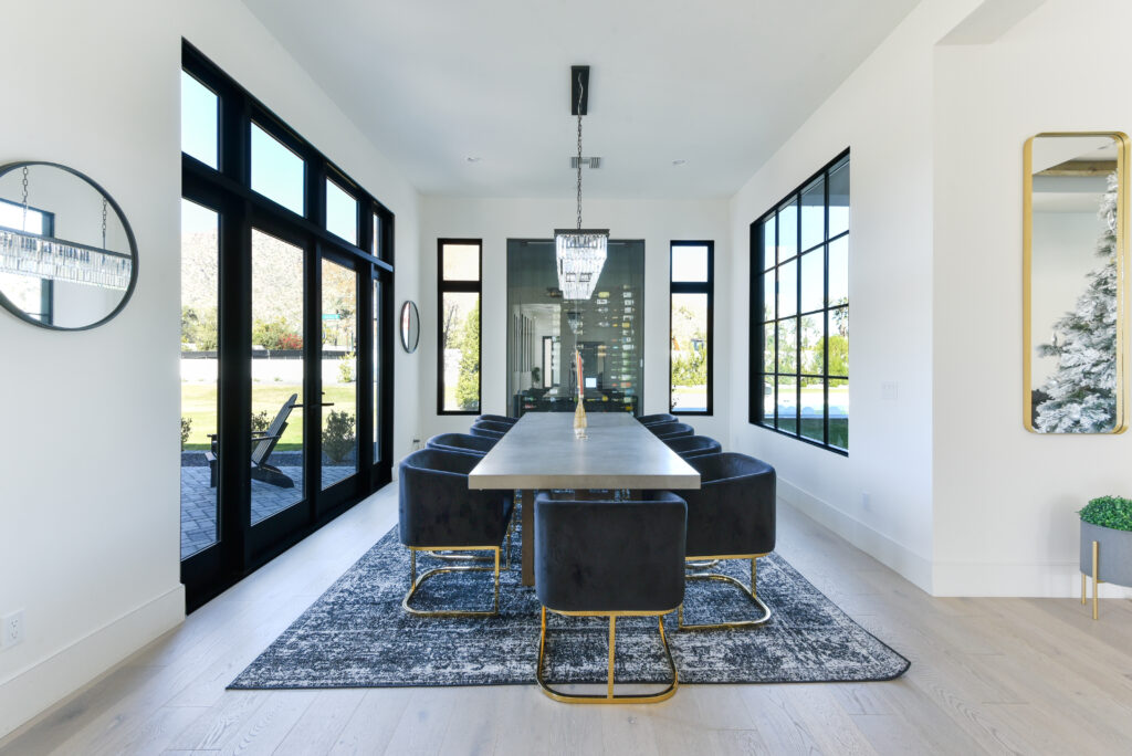 Dining room with a long concrete table and dark velvet chairs on gold bases over a patterned rug on light hardwood floors, lit by black-framed French doors and grid windows opening to the patio, lawn and mountains, with a glass-enclosed wine display and a crystal chandelier behind