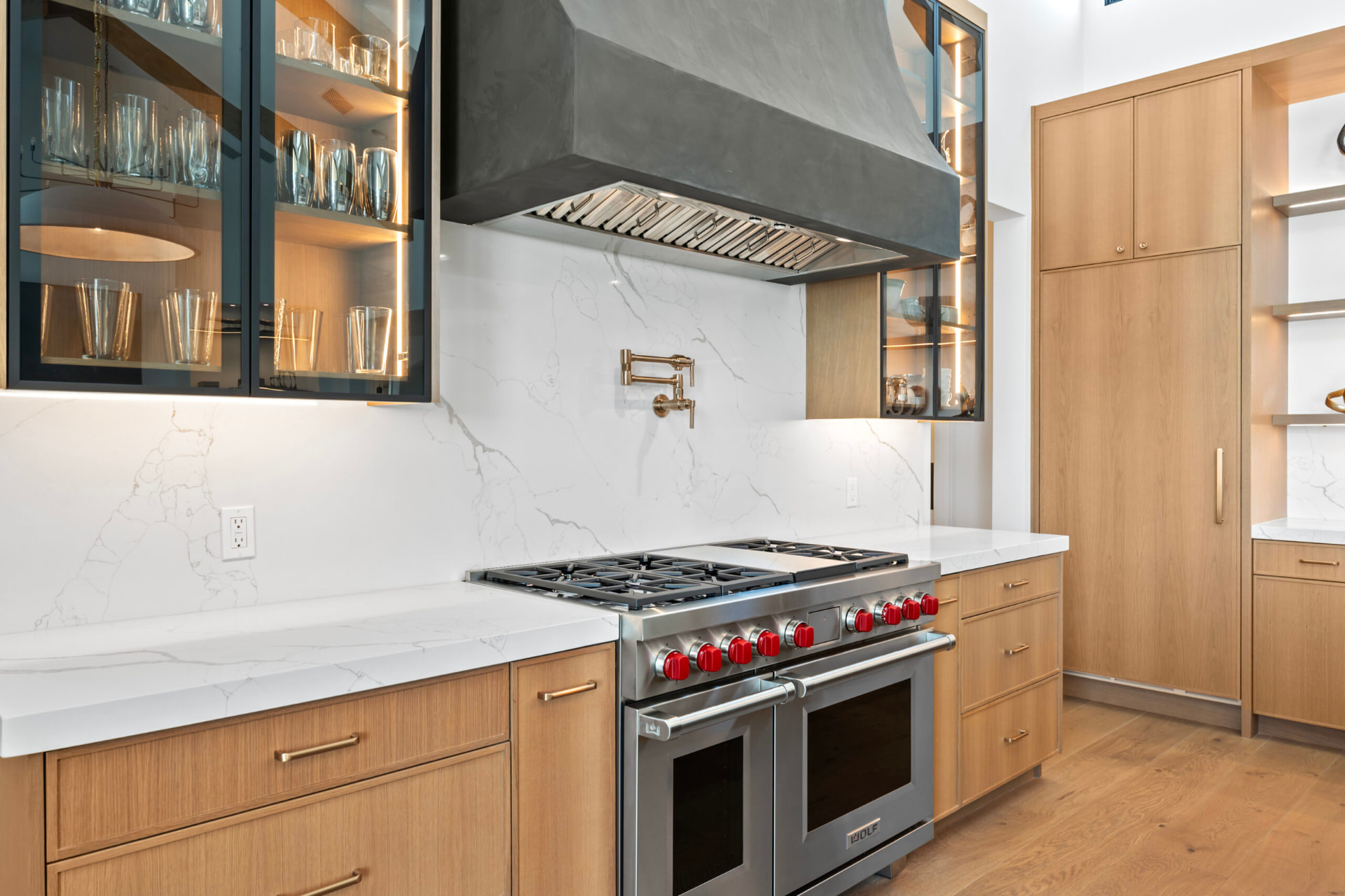 Kitchen detail showing a stainless steel range with red knobs beneath a wide charcoal-gray hood and a brass pot filler, with light natural wood cabinetry, white marble counters and backsplash, and interior-lit glass-front display cabinets