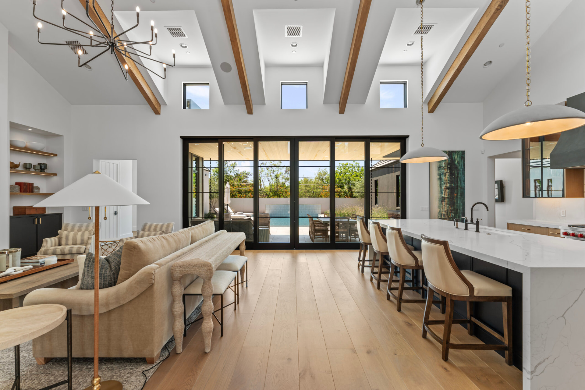 Open-concept great room and kitchen beneath a high vaulted ceiling with exposed wood beams and clerestory windows, lit by full-height black-trimmed sliding glass doors onto the pool patio, with light oak floors, a black island topped in white waterfall marble with leather barstools, and a neutral beige living area beyond