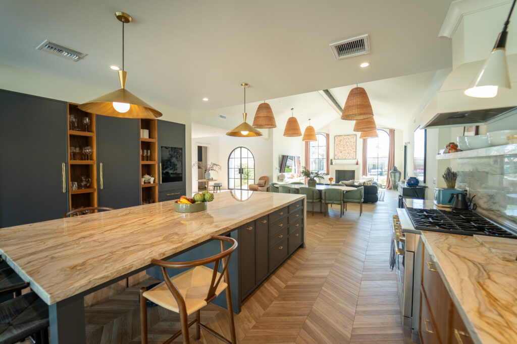 Kitchen with a large beige marble-topped island, dark grey cabinetry, brass cone pendant lights and light herringbone wood floors, opening to the dining and living space